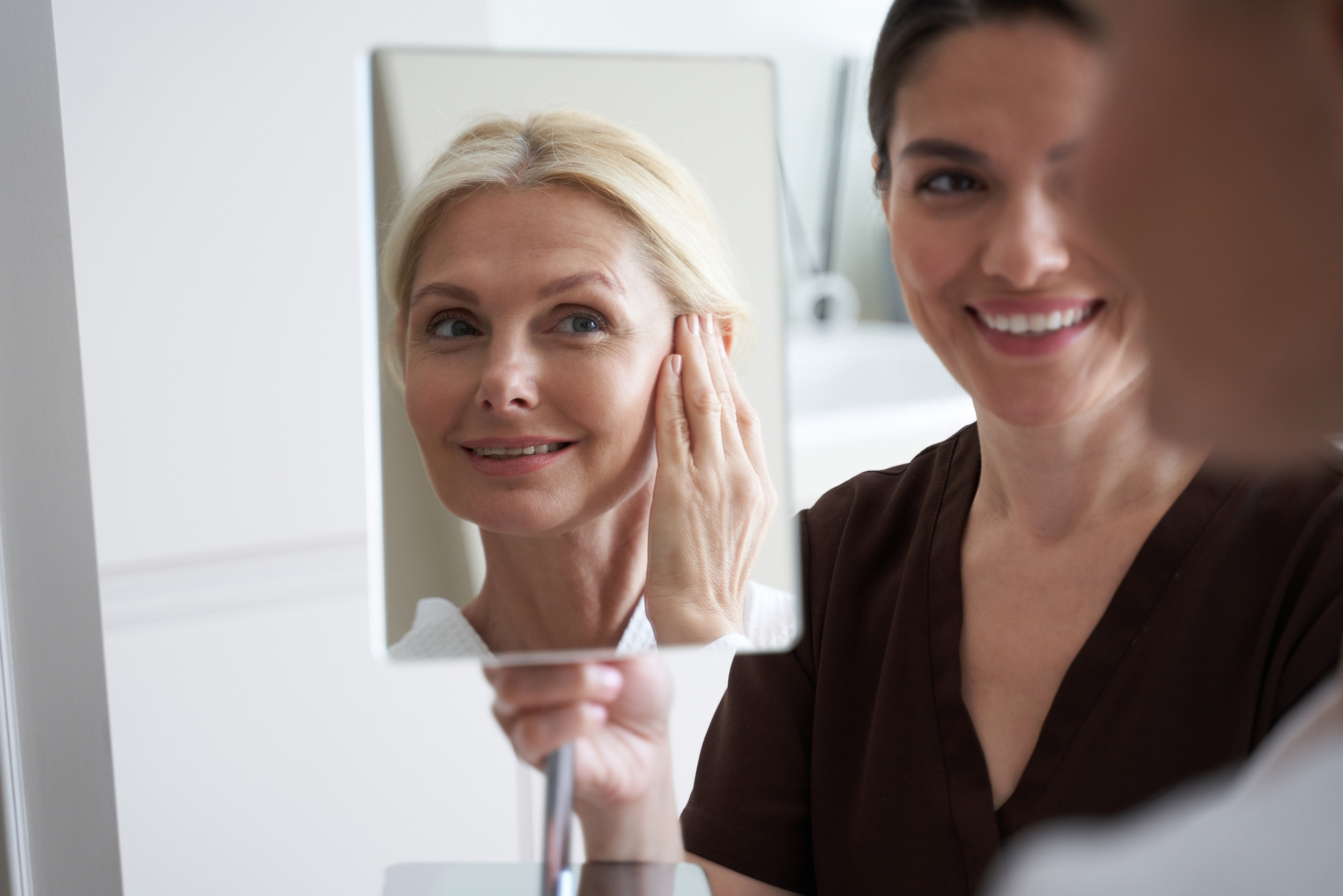 Smiling attractive lady enjoying after rejuvenating facial treatments Happy adult woman looking at her reflection in the mirror and sitting in the office next to her beautician